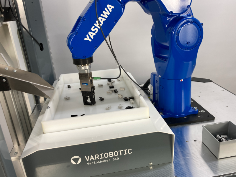 Ein blauer Yaskawa-Industrieroboter greift Kleinteile aus einer weißen VarioShaker 540 Sortiereinheit von Variobotic.